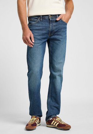 Jeans in denim blu scuro con taglio dritto, cuciture a contrasto, cinque tasche e chiusura con bottone, abbinati a sneakers marroni e gialle.