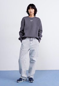 Grijze sweatshirt met een wit Nike-logo, gecombineerd met lichtgrijze baggy jeans en zwarte sportschoenen met zilveren accenten.