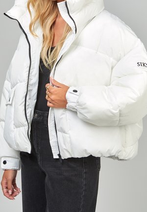 Femme portant une doudoune blanche avec une fermeture éclair noire et des boutons, un haut noir et un jean taille haute foncé, tenant la veste près de la fermeture éclair.
