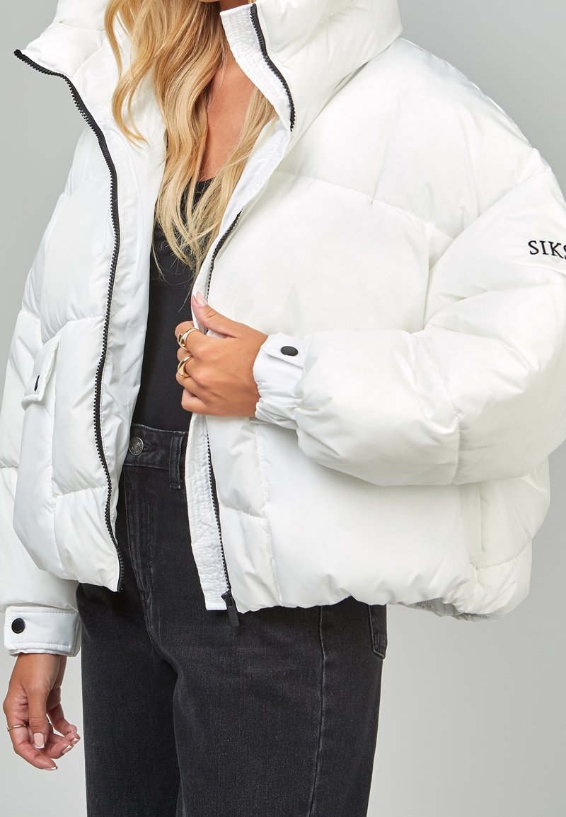 Femme portant une doudoune blanche avec une fermeture éclair noire et des boutons, un haut noir et un jean taille haute foncé, tenant la veste près de la fermeture éclair.