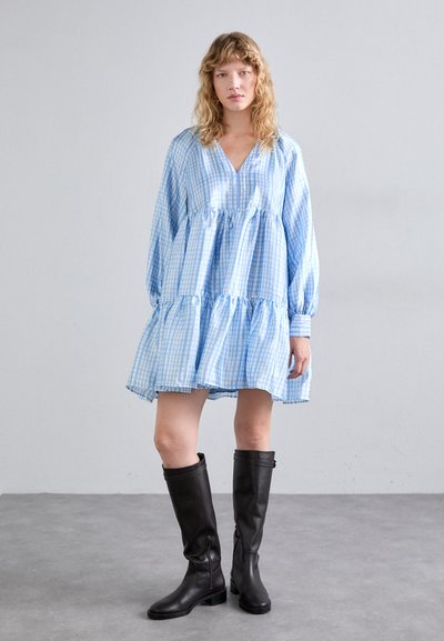 Robe à carreaux bleus et blancs avec un ourlet en étages, des manches longues et un décolleté en V. Associée à des bottes noires en cuir hautes. Texture lisse.