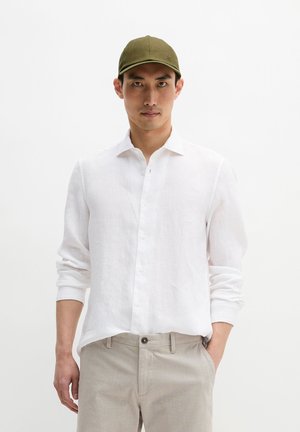 Młody mężczyzna w oliwkowej czapce, białej koszuli z podwiniętymi rękawami i beżowych spodniach, stojący na białym, jednolitym tle.