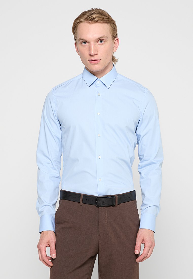 Uomo che indossa una camicia dress blu chiaro infilata in pantaloni marroni con una cintura nera, in piedi contro uno sfondo chiaro e semplice.