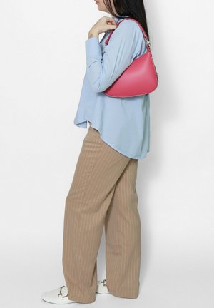 Donna con camicetta celeste e pantaloni beige a righe sottili che tiene una borsa rossa di media dimensione sulla spalla.