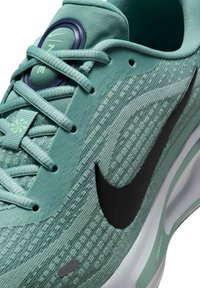 Zielony but sportowy z teksturowaną siateczką, czarnym logo Nike i okrągłą podeszwą. Zawiera okrągłe zapięcie sznurowadeł i wzorzyste akcenty.