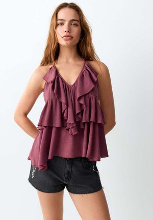 Femme aux longs cheveux portant un haut violet sans manches à volants et un short en jean noir sur un fond blanc uni.