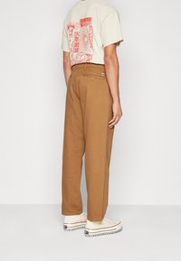 Bruna vida benbyxor med en slät struktur, med bakfickor och bälteshällor. Bärs med en beige grafisk t-shirt och vita sneakers.