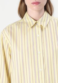 Camicia gialla con bottoni, a righe verticali sottili rosse, dotata di colletto standard e bottoni bianchi sul fronte. Tessuto morbido e liscio.