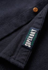 Tissu bleu marine foncé avec une étiquette Superdry cousue en texte blanc et vert près d'un bouton en bois sur un vêtement.