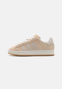 Beige suede sneakers with textured side panels, three subtle stripes, round laces, and a white midsole over a gum rubber outsole.