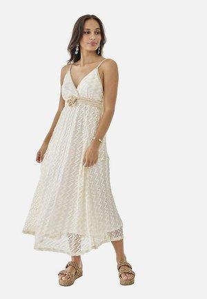 Mujer con un vestido largo sin mangas de encaje color crema con un cinturón de flores, pendientes turquesas, pulsera y sandalias de plataforma.