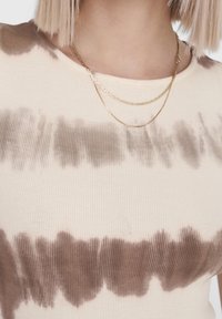 Kremowy, prążkowany top z brązowym wzorem tie-dye, posiadający szeroki dekolt i nawarstwione złote naszyjniki. Materiał jest miękki i dopasowany.