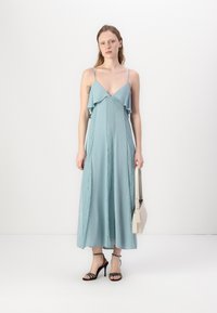 Svetlo modra maxi obleka s plastastim volanom, tankimi naramnicami in čipkastimi detajli, v kombinaciji s črnimi sandalmi na peto in bež torbico.