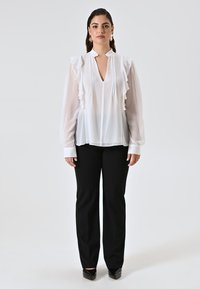 Donna in piedi, rivolta in avanti, che indossa una camicia bianca con volant, pantaloni neri e scarpe con punta nera, su uno sfondo chiaro e semplice.