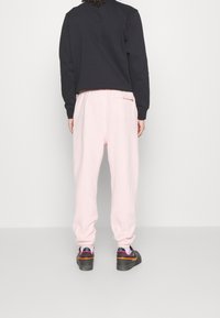 Person som bär ljusrosa sweatpants, svart sweatshirt och svarta sneakers med orange och lila detaljer, stående mot en enhetlig ljus bakgrund.