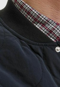 Zwarte gewatteerde jas met kraag en een getextureerde elastische band, voorzien van een rood-wit geruit binnenfutter en een zilveren drukknop.