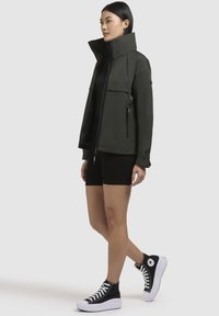 Giacca verde scuro, con zip frontale, colletto alto, due tasche laterali, indossata sopra un body nero e pantaloni corti neri, abbinata a sneakers alte nere.