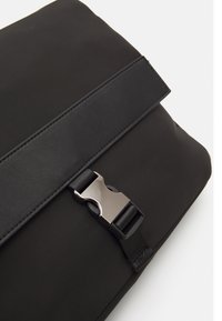 Borsa a tracolla nera realizzata in tessuto resistente, con una texture liscia, una larga patta e una chiusura con gancio metallico.