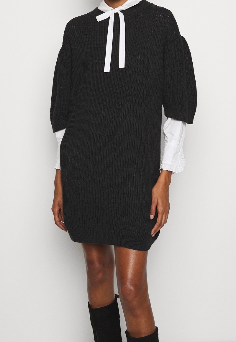 Abito nero in maglia con silhouette rilassata, maniche a sbuffo e colletto in raso bianco. La texture è a coste con un finish morbido.
