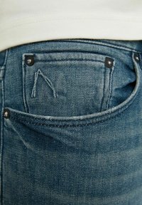 Gros plan sur la poche arrière d'un jean en denim bleu avec des détails de couture blanche et des rivets noirs, porté avec une chemise blanche.