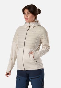 Femme portant une veste matelassée beige clair avec capuche et un pantalon bleu marine, debout avec une main dans la poche, regardant sur le côté.