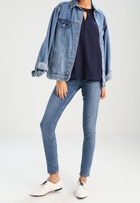 Veste en denim, blouse marine, jean skinny et chaussures blanches à lacets. La veste est bleu clair avec des poches ; la blouse présente un détail en forme de trou de serrure.