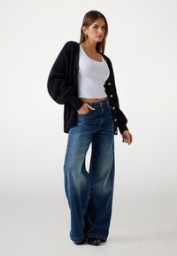 Black ribbed cardigan with large buttons, white fitted tank top, and wide-leg distressed blue jeans. Smooth texture, slightly faded color.