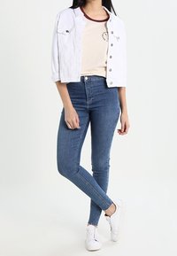 Vit croppad denimjacka, beige T-shirt med vinröda accenter och blå slim fit jeans; ihop med vita sneakers.