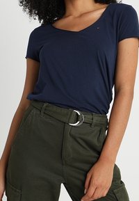 T-shirt bleu marine à manches courtes avec un col en V, associé à un pantalon taille haute en olive texturé, doté d'une boucle de ceinture circulaire en argent.