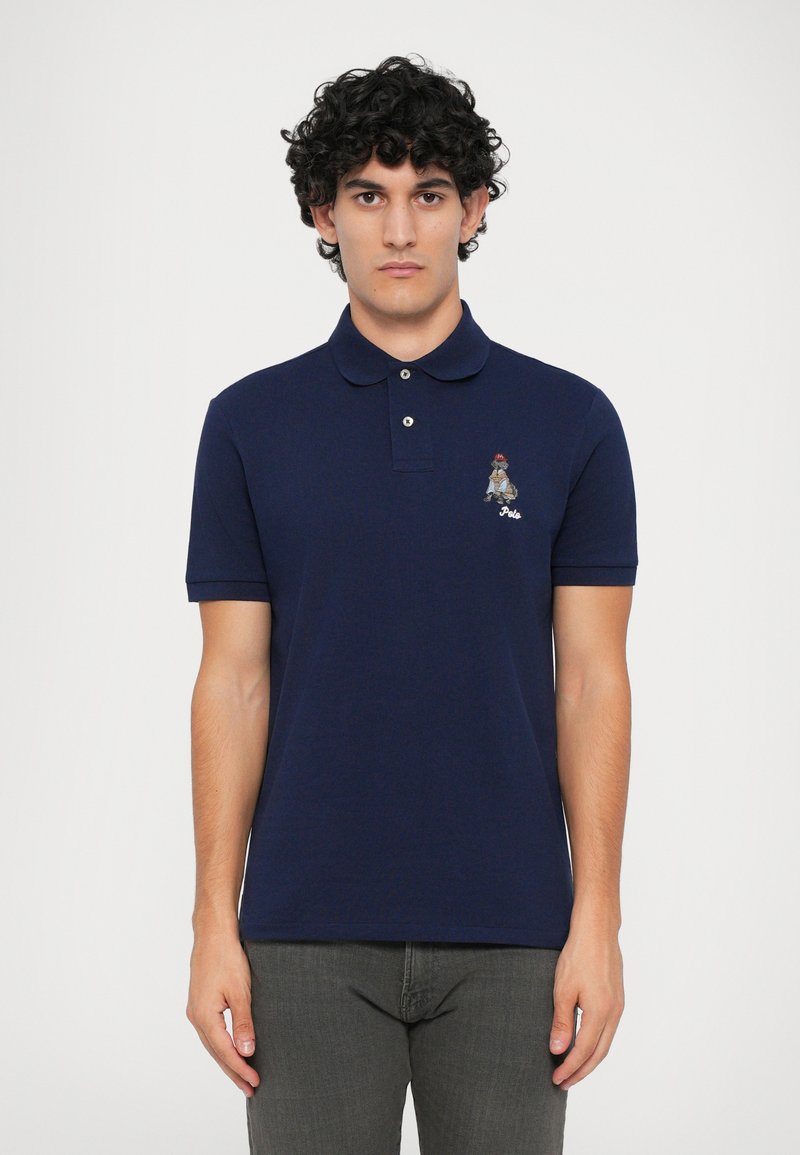 Jovem com cabelo encaracolado veste uma camisa polo azul escura com um pequeno logótipo de urso bordado no lado esquerdo do peito, combinada com calças cinzentas.