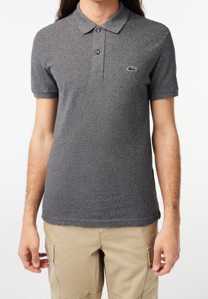 Mand iført en grå kortærmet poloshirt med et broderet krokodillelogo på brystet og beige cargobukser.