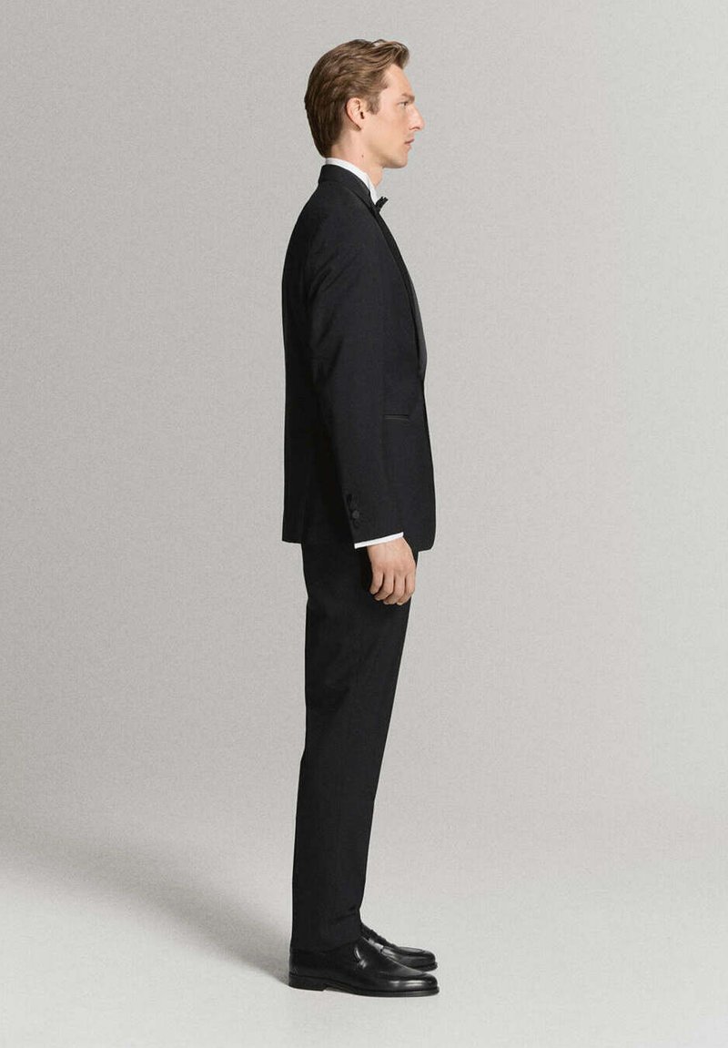 Hombre de perfil vestido con un esmoquin negro con pajarita y zapatos de vestir negros sobre un fondo liso y claro.