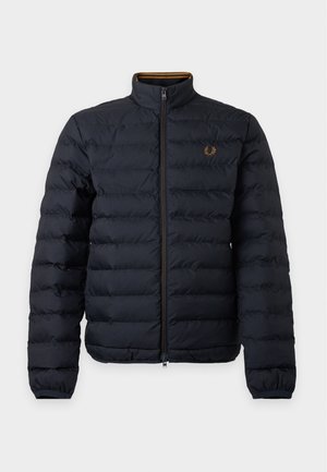 Homme debout portant une veste matelassée noire, un pantalon blanc et des chaussures noires sur un fond clair uni.