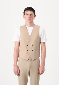 Gilet beige doppiopetto con bottoni neri, progettato in una vestibilità su misura, abbinato a una semplice t-shirt bianca e pantaloni coordinati.