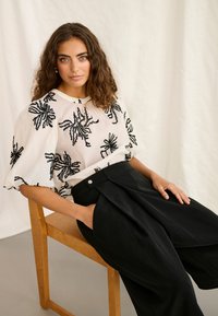 Femme aux cheveux bruns bouclés assise sur une chaise en bois, portant un chemisier blanc avec une broderie florale noire et un pantalon noir taille haute à jambes larges.