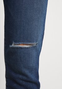 Jeans de mezclilla azul oscuro con una rodilla desgastada que presenta un pequeño desgarro horizontal, bordes deshilachados y una textura suave en toda la prenda.