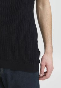 Granatowy sweter w prążki o fakturowanym wzorze, dopasowany w talii i z akcentem na bocznej metce. Materiał wydaje się miękki i elastyczny. Ręka spoczywa w pobliżu.