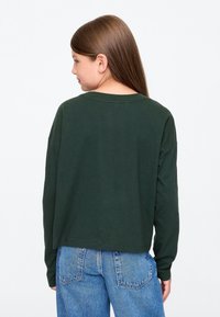 Sötétzöld, hosszú ujjú ing, laza szabással és széles nyakkivágással, világoskék, magas derekú farmernadrággal párosítva. Lágy anyagtextúra.