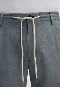 Leichte, graue Hose aus strukturiertem Stoff. Mit elastischem Bund, weißem Kordelzug und einem kleinen Knopfdetail am Bund.