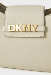 Bežinė rankinė su tekstūruotu paviršiumi, kuriame yra auksinės metalinės „DKNY“ logotipo ir užsegimo su atitinkama petneša.