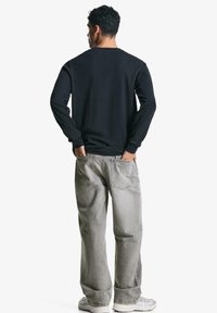 Black long-sleeve shirt with round neck, paired with light gray loose-fit jeans. The person stands with hands in pockets, facing away.