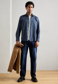 Σκούρο μπλε καρό πουκάμισο, λευκή μπλούζα, μπλε τζην, κρατώντας ένα καφέ μπουφάν, μαύρα παπούτσια, στέκεται σε ξύλινο πάτωμα με ουδέτερο φόντο.