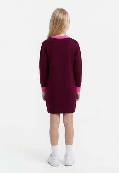 Dieťa s dlhými blond vlasmi stojí otočené chrbtom, má na sebe bordové šaty s ružovými manžetami a golierikom, biele ponožky a biele tenisky.