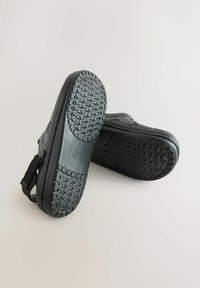 Schwarze wasserdichte Slipper mit strukturierten Gummisohlen und verstellbaren Fersenriemen, mit nach außen gerichteten Sohlen auf einem schlichten Hintergrund gezeigt.