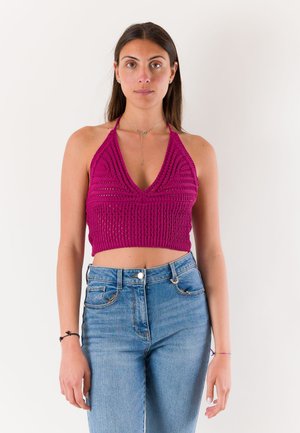 Donna che indossa un top corto magenta lavorato a maglia con collo halter e jeans blu a vita alta, in piedi davanti a uno sfondo bianco uniforme.