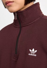 Maronenfarbener Pullover mit Viertelreißverschluss aus strukturiertem Stoff; verfügt über einen schwarzen Reißverschluss und ein weißes, gesticktes Adidas-Logo auf der linken Brust.