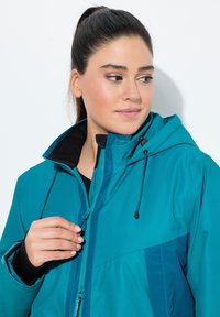 Teal zip-jacka med huva, med en svart fodrad krage, justerbara dragsnören och kontrasterande färgpaneler. Slät, vattentålig tyg.