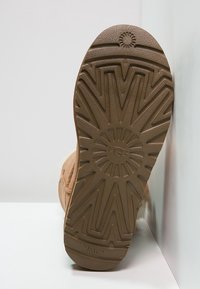 Suela de goma marrón con un patrón en zigzag texturizado y un logotipo circular. La parte superior muestra un material suave en un tono neutro.