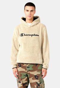 Sudadera de forro polar beige con un logo negro de "Champion", que presenta capucha y una textura suave. Combinada con pantalones de camuflaje, mostrando un ajuste relajado.