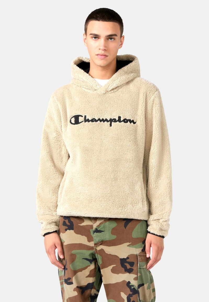 Sudadera de forro polar beige con un logo negro de "Champion", que presenta capucha y una textura suave. Combinada con pantalones de camuflaje, mostrando un ajuste relajado.
